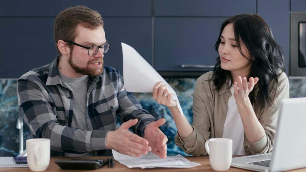 Two people sit at a table with a laptop, mugs, and several papers during a discussion.