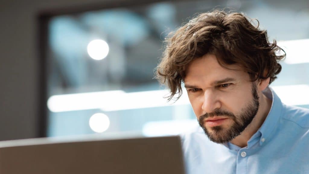 A man with curly brown hair and a beard looks down at a laptop screen.