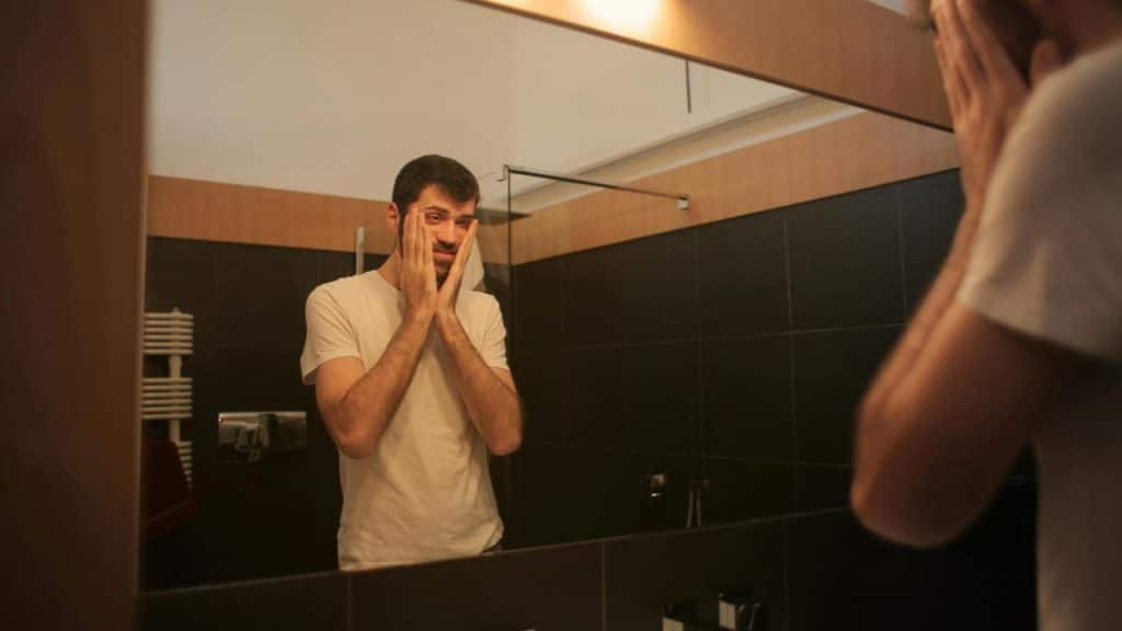 A man touching his face while looking in a bathroom mirror.