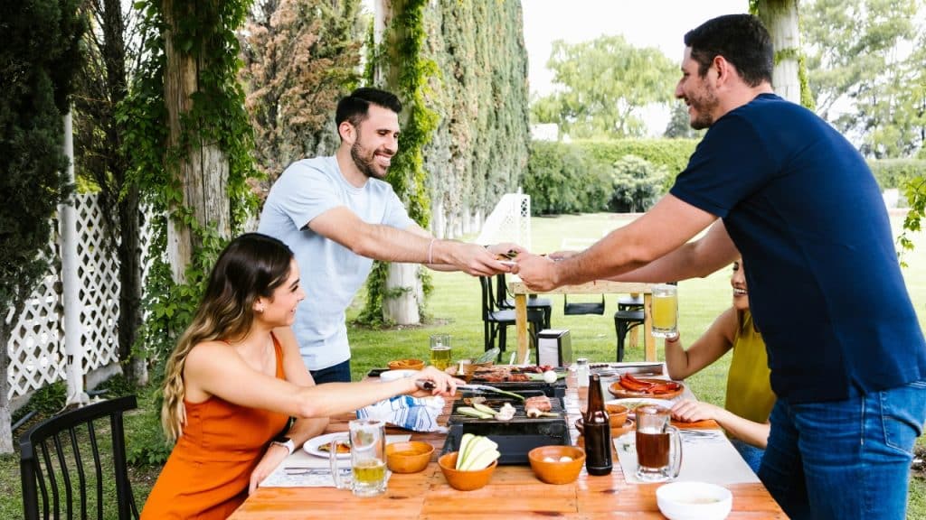 A group of people interact around a grill and outdoor dining table.