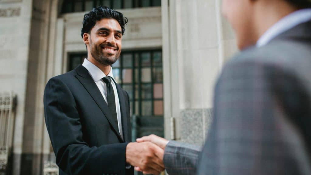 A man in a dark suit smiles while shaking hands with someone in a blazer.