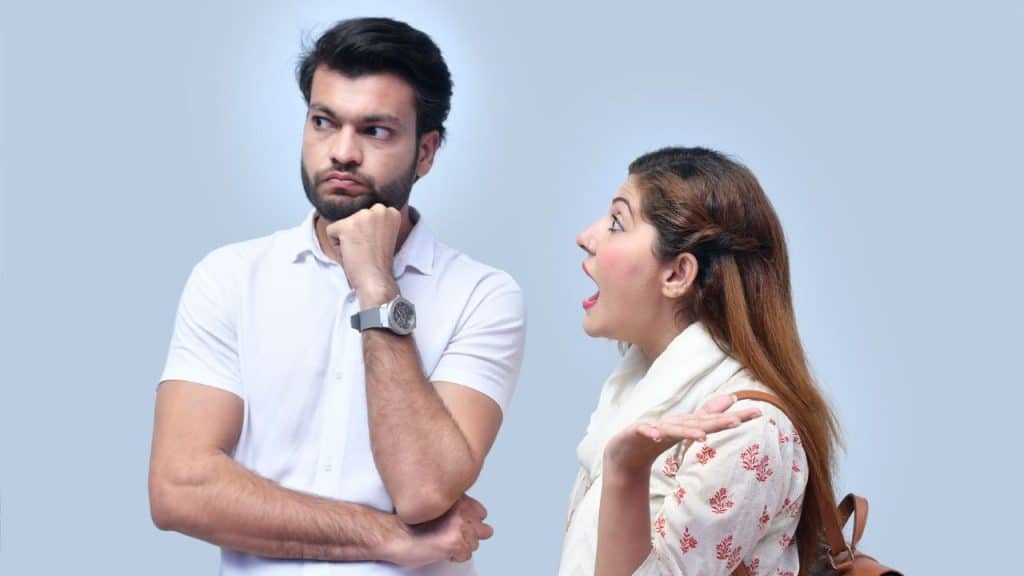 A woman with her hand raised speaks to a man who is looking away thoughtfully.