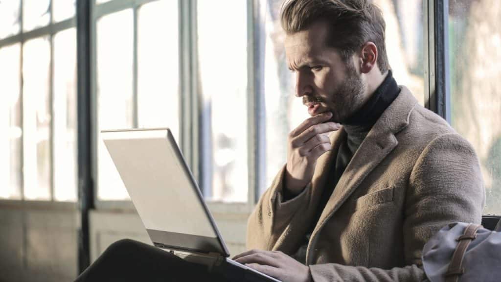 A man in a tan coat sits with his hand on his chin, looking at a laptop.