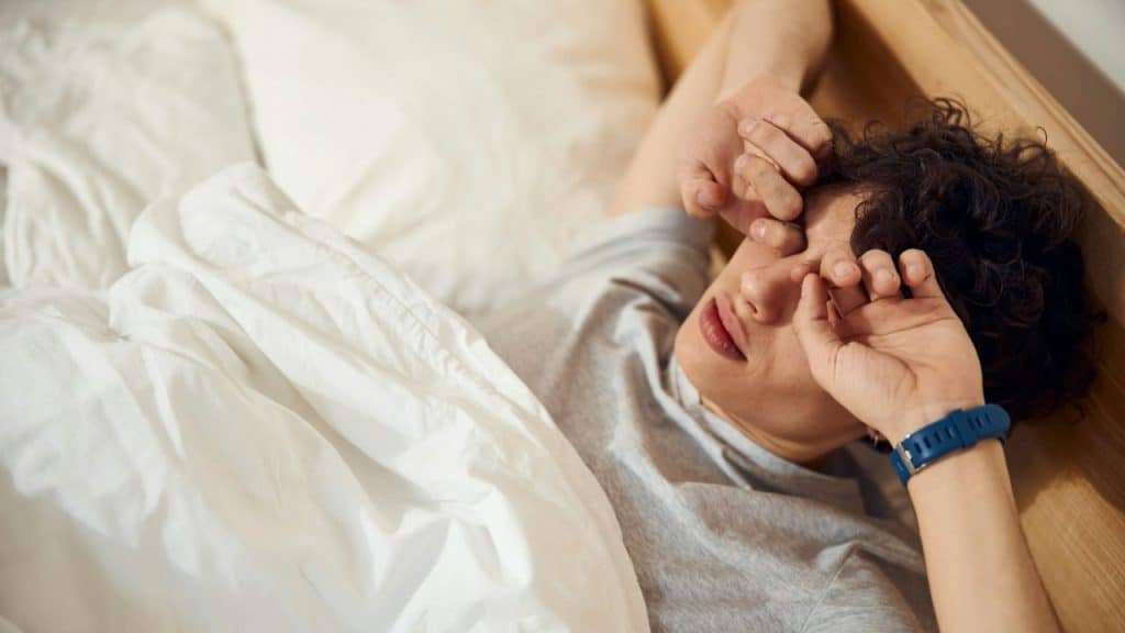 A person lies in bed under a white blanket and rubs their eyes with their hands.