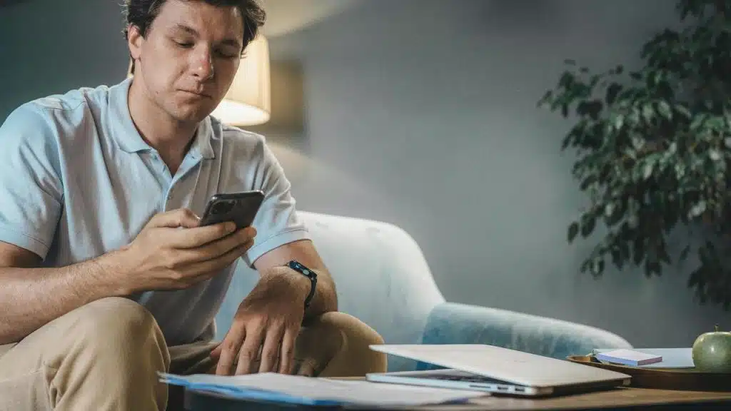 A man sitting on a couch looking at his phone beside a table with papers and a laptop.