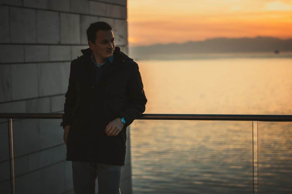A man leaning against a rail by the sea at sunset and looking to the side.