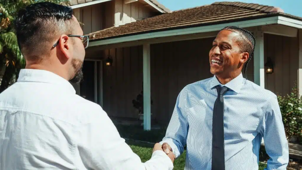 Two men shake hands outdoors in front of a house, one smiling and wearing a tie.