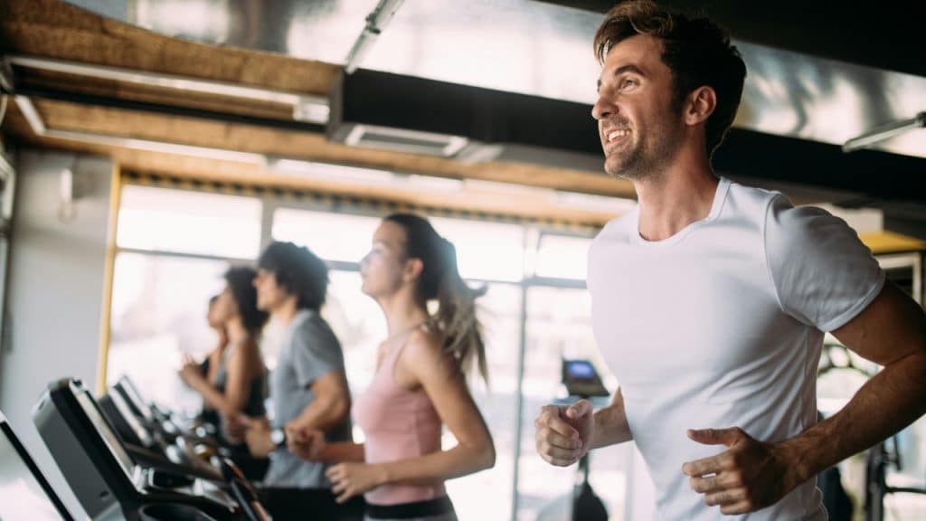 People run on treadmills in a gym while a man in front smiles.
