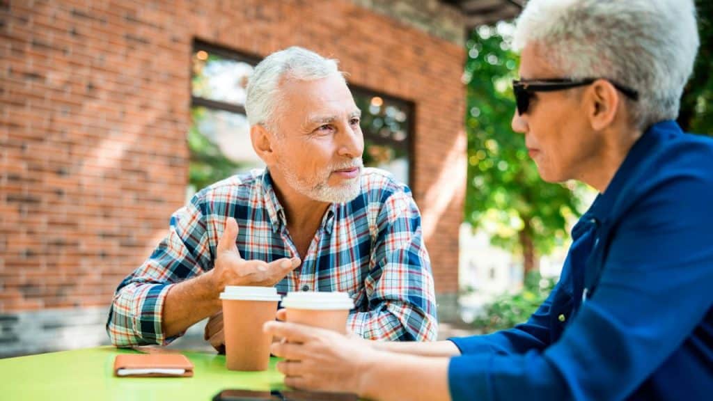 A man in a plaid shirt gestures while talking to a woman at a table.