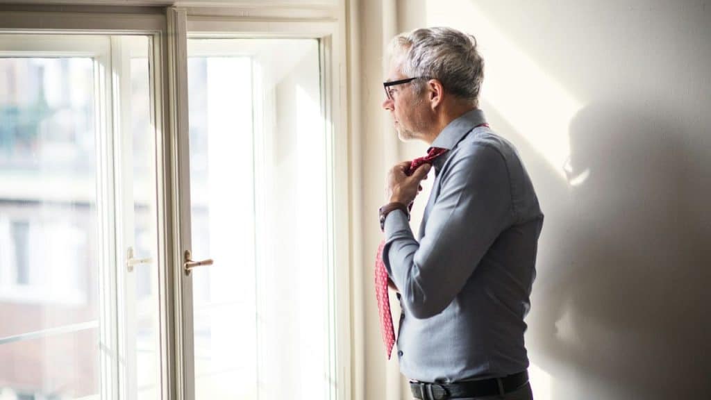 A man in a dress shirt adjusts his red necktie while looking out a window.