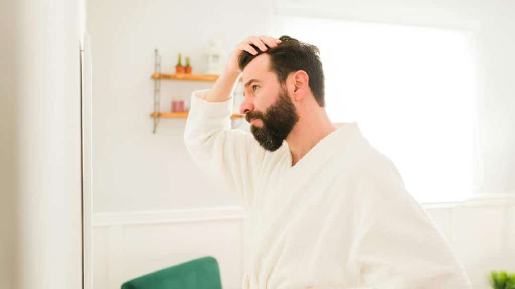 A bearded man touching his hair while looking in a mirror.