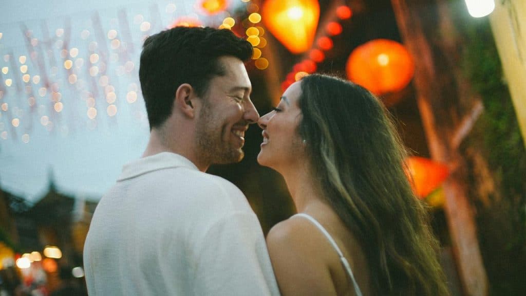 A couple smiling closely under glowing lantern lights at night.