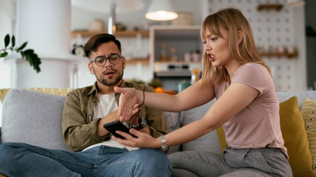 A woman points toward a smartphone screen held by a man sitting on a sofa.