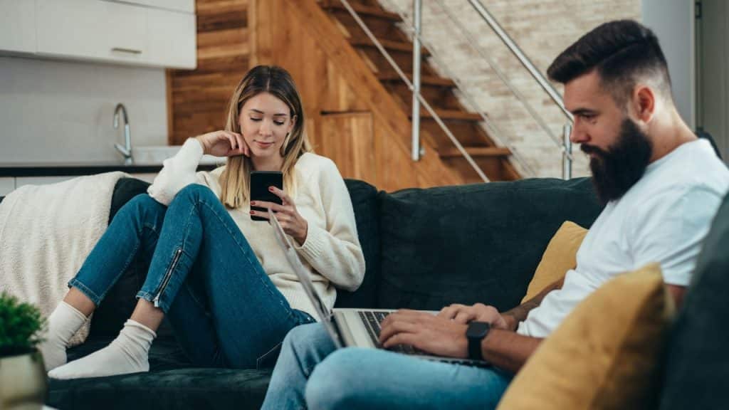 A woman uses her phone while a man works on a laptop nearby on furniture.
