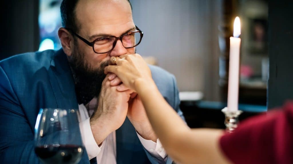A bearded man in a suit holds his partner's hands at a table.