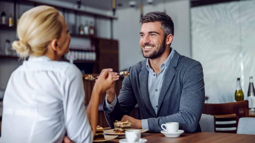 A smiling man in a blazer sits at a table across from a blonde woman.