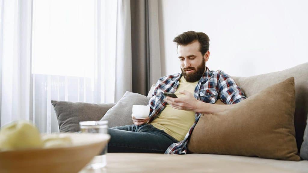 A man sitting on a couch holding a cup and looking at his phone.