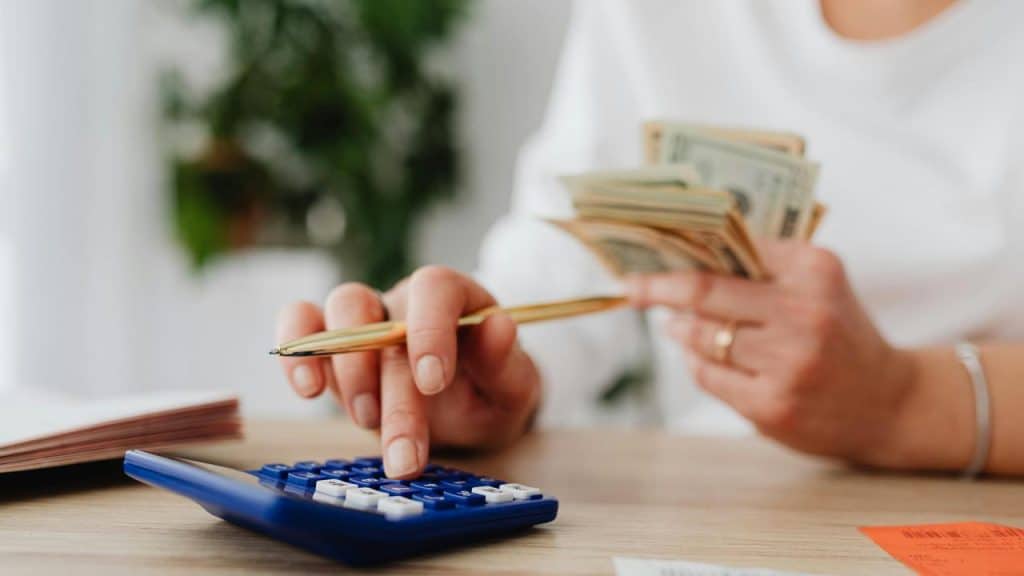 A person using a calculator while holding cash and a pen at a desk.