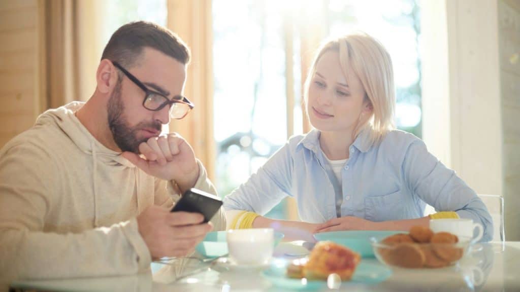 A man looks at a phone while a woman watches him at a sunlit breakfast table.