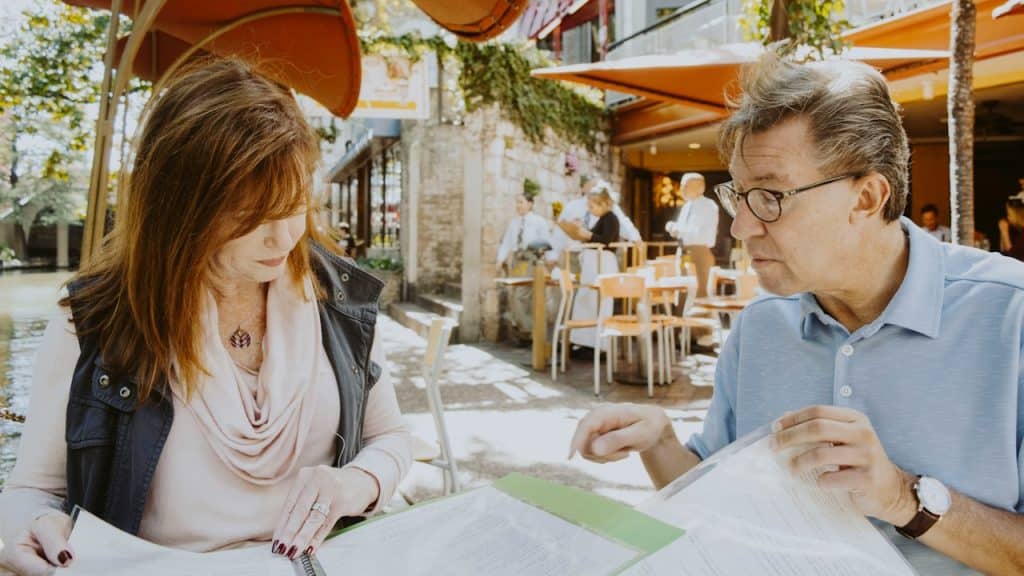 A man and a woman are sitting down near a table while reading financial documents outside a cafe during the daytime.