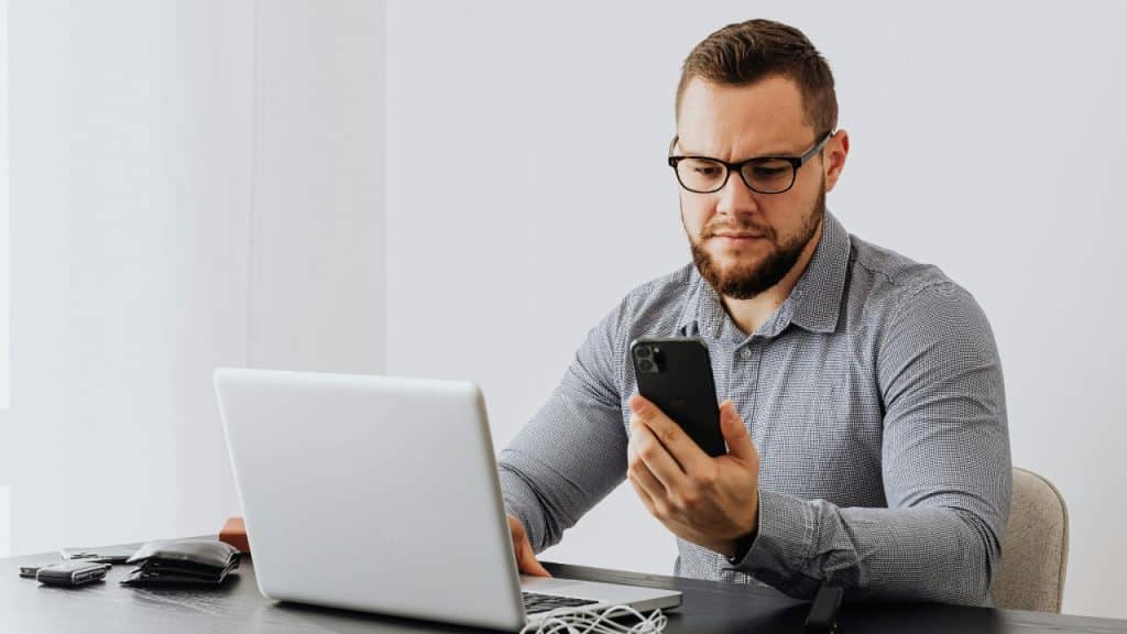 A man wearing glasses sits at a desk, looking at his smartphone and laptop.