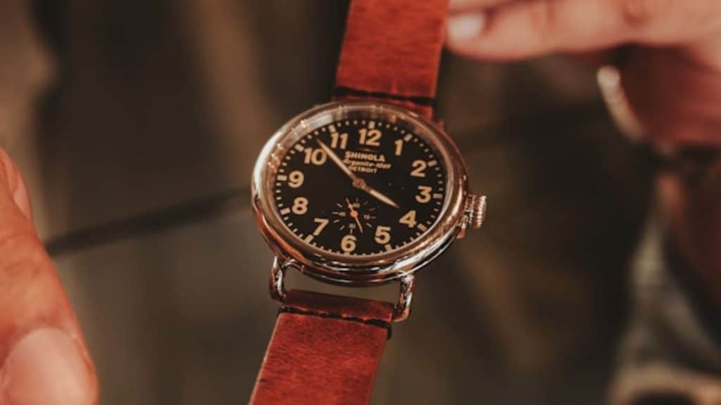 Hands hold a Shinola wristwatch with a black face and brown leather strap.