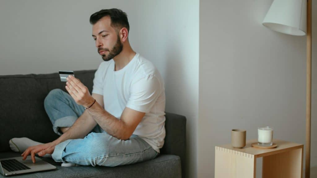 A man sits on a couch using a laptop while holding a credit card.