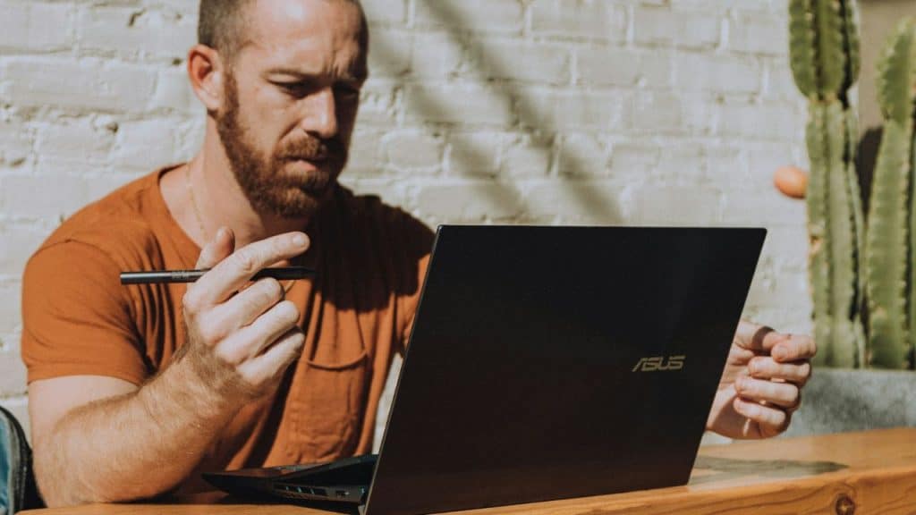 A man with a beard and orange shirt works on an ASUS laptop.
