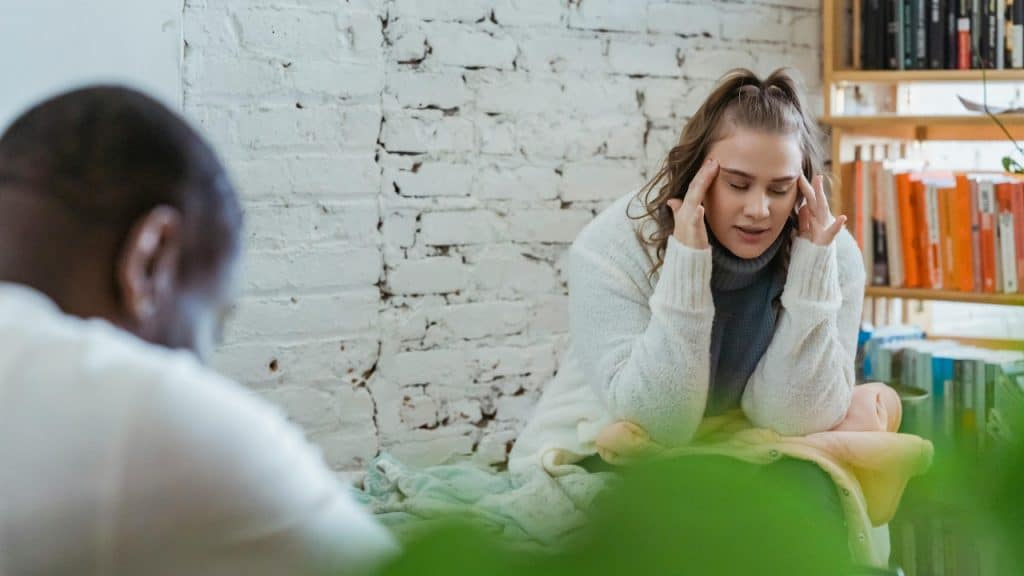 A woman sits holding her head while a man sits blurred in foreground.