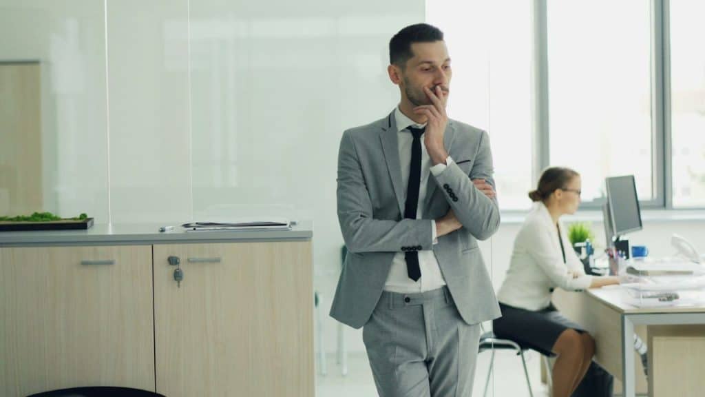 A man in a grey suit stands in an office with his hand on his chin.