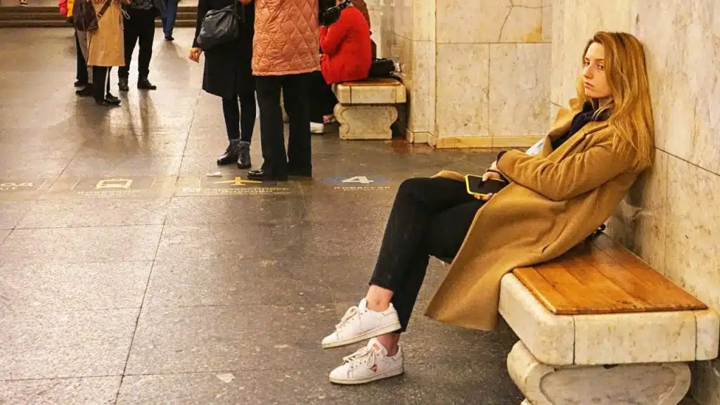 A woman sits alone on a bench in a station while others stand nearby.