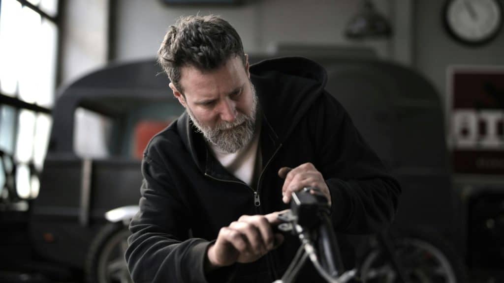 A bearded man in a black hoodie works on the handlebars of a motorcycle.