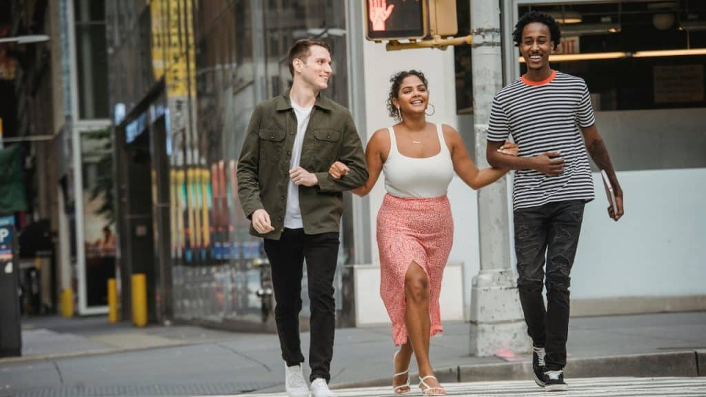Three people walk together across a city street, with the woman in the center linking arms.