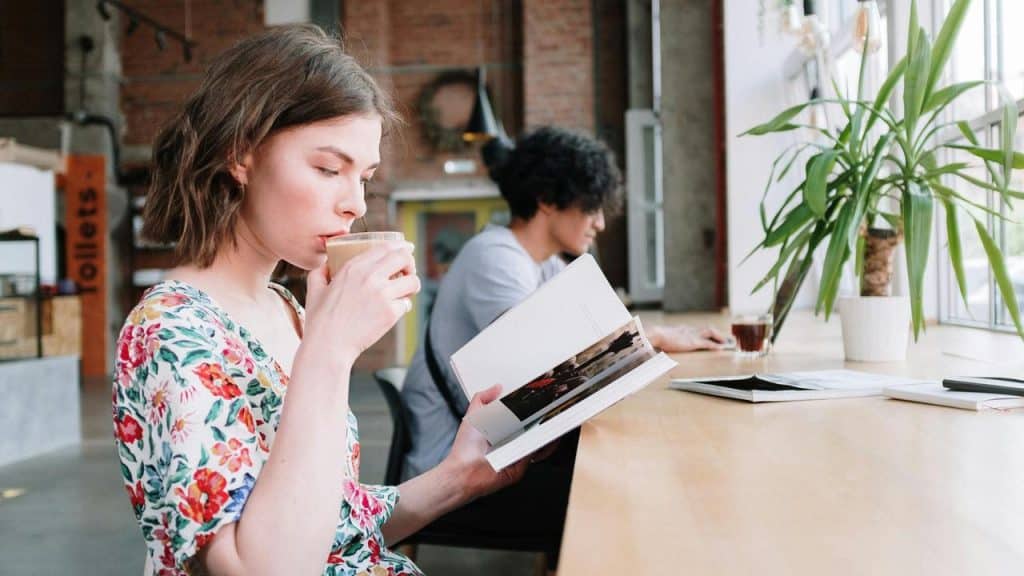 A woman reading a book and drinking coffee at a café table.