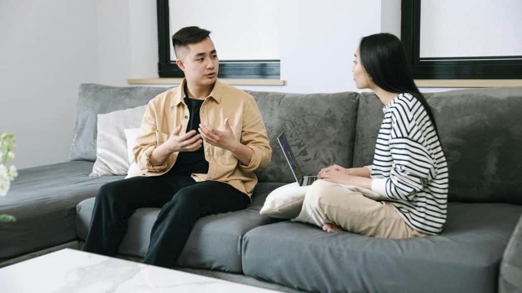 A man and a woman sitting on a couch, talking while using a laptop.