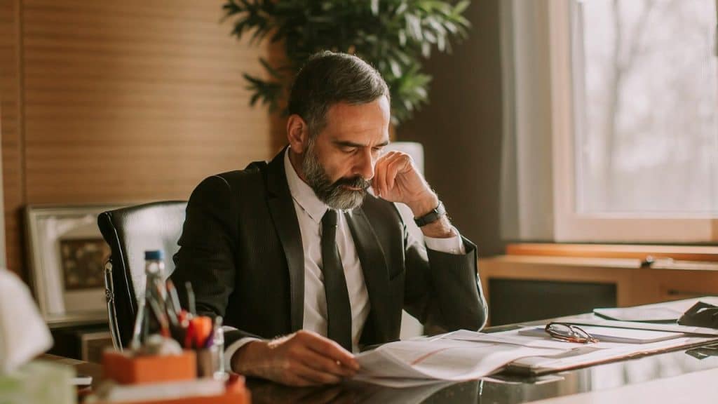 A bearded man in a suit sits at a desk looking at some documents.