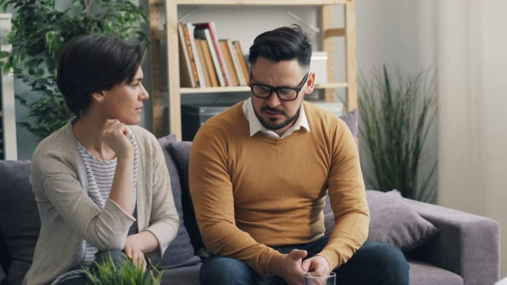 A woman sits beside a man on a couch, looking at him while he looks down.