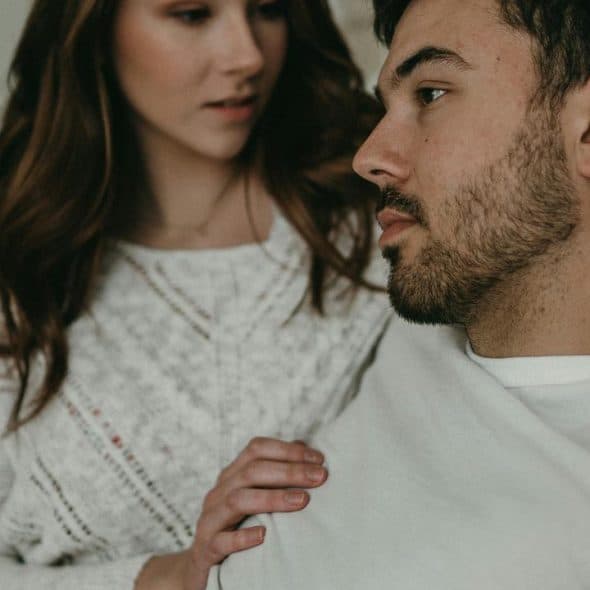 A woman gently touching a seated man’s shoulder as they share a quiet moment indoors.