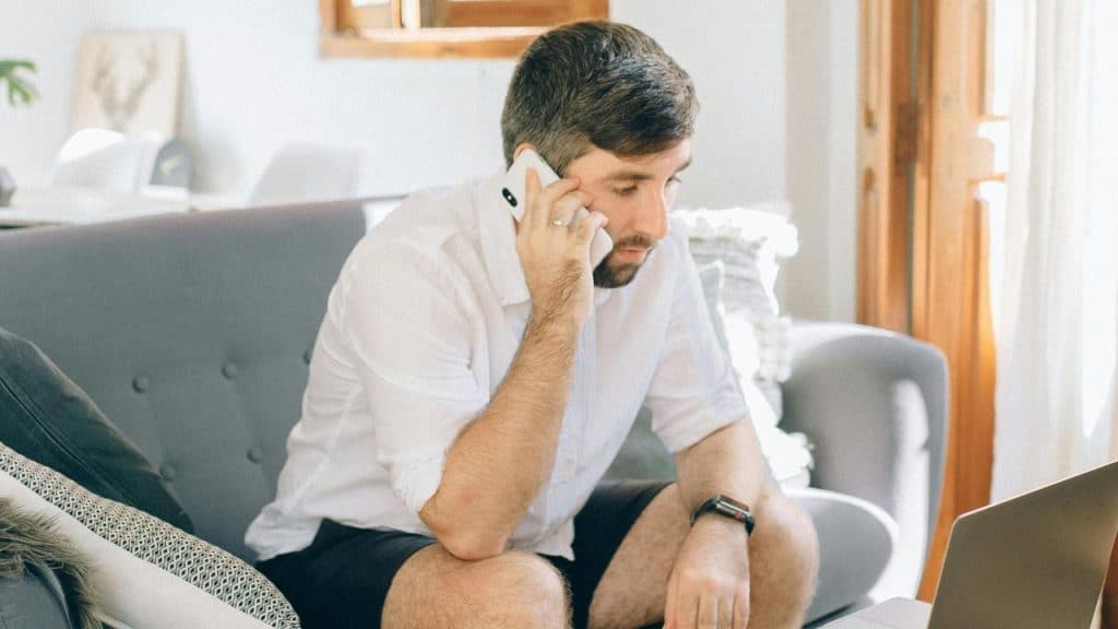 A man sits on a couch talking on his phone near a laptop.