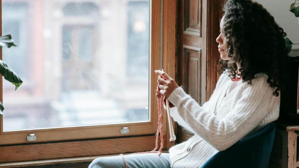 A woman sitting by a window knitting with red yarn.