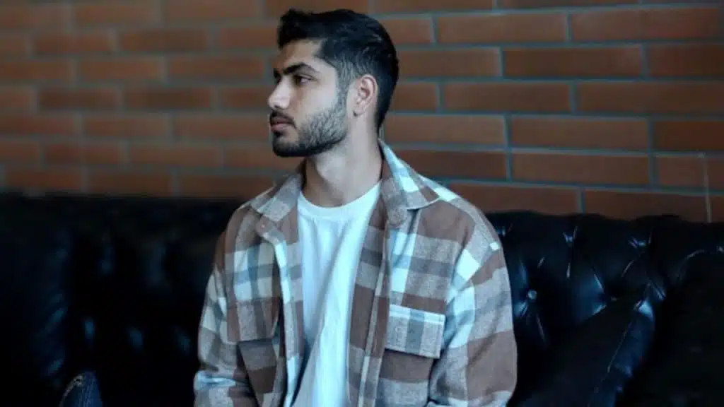 A bearded man in a plaid overshirt sits on a black leather sofa.