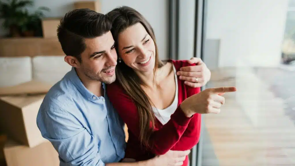 A man embraces a woman from behind while she points toward an open window.