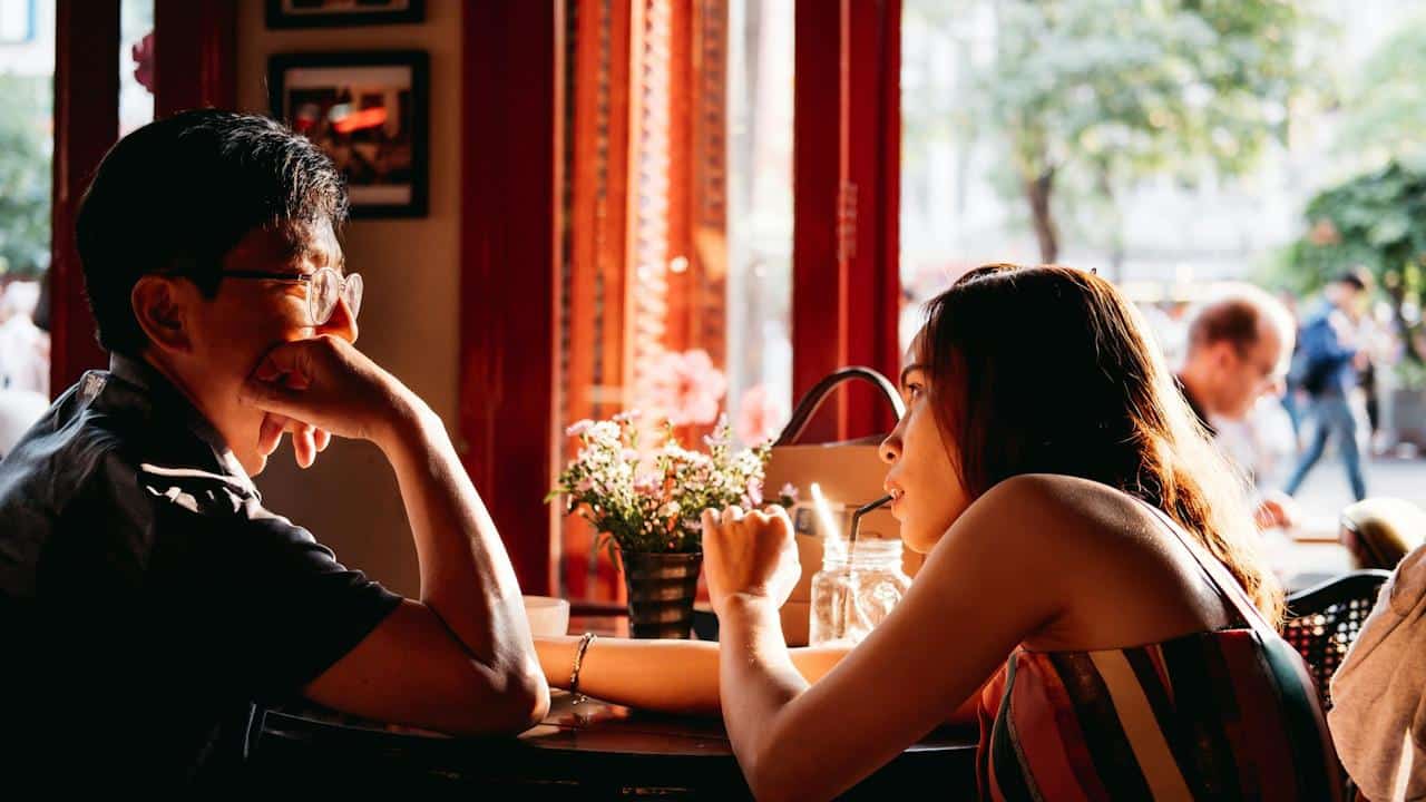 Two people sit across from each other at a small café table by a window, engaged in conversation.