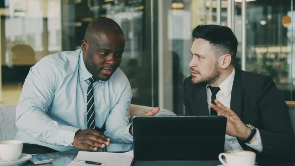 Two men in business attire sit at a table with a laptop, gesturing while speaking.
