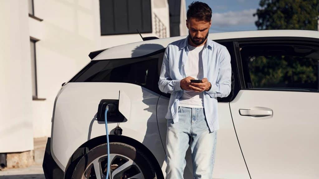 A man checking his phone beside a charging electric car.