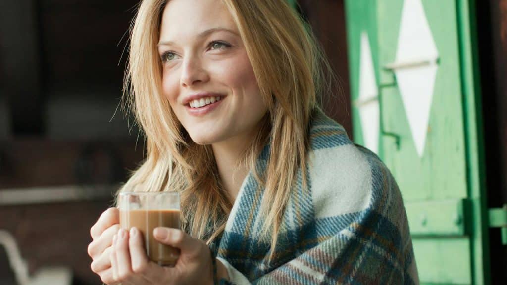 A smiling woman holding a warm drink near a window.