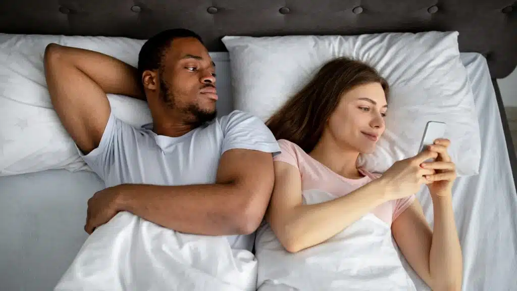 A man looks at a woman lying in bed, who is focused on her phone.