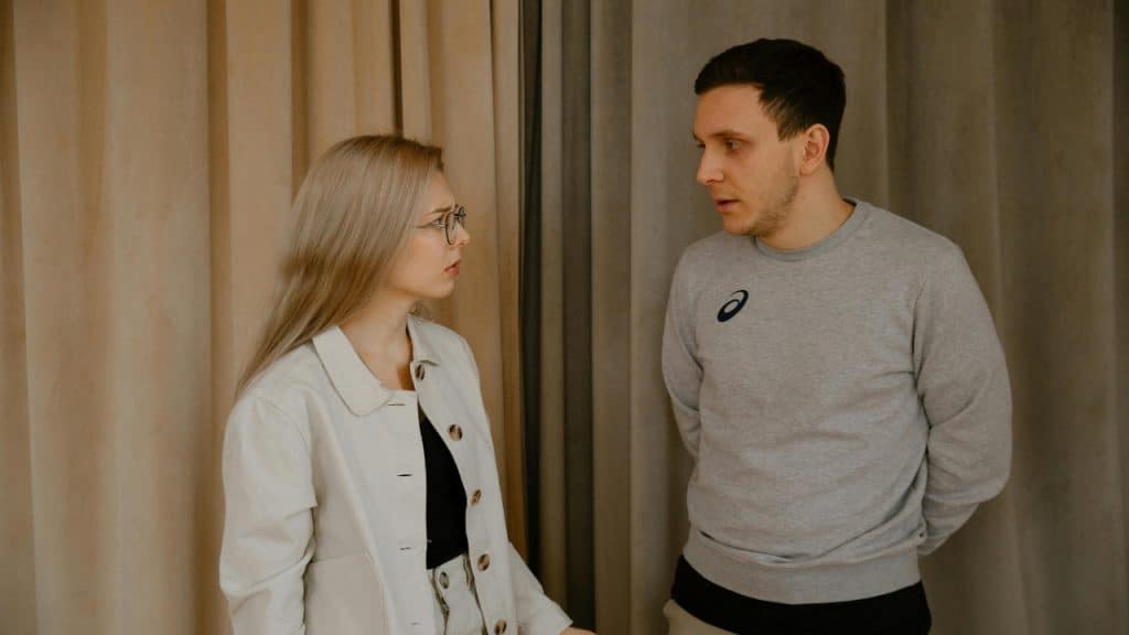 A man in a grey sweatshirt and a woman in a white jacket face each other.