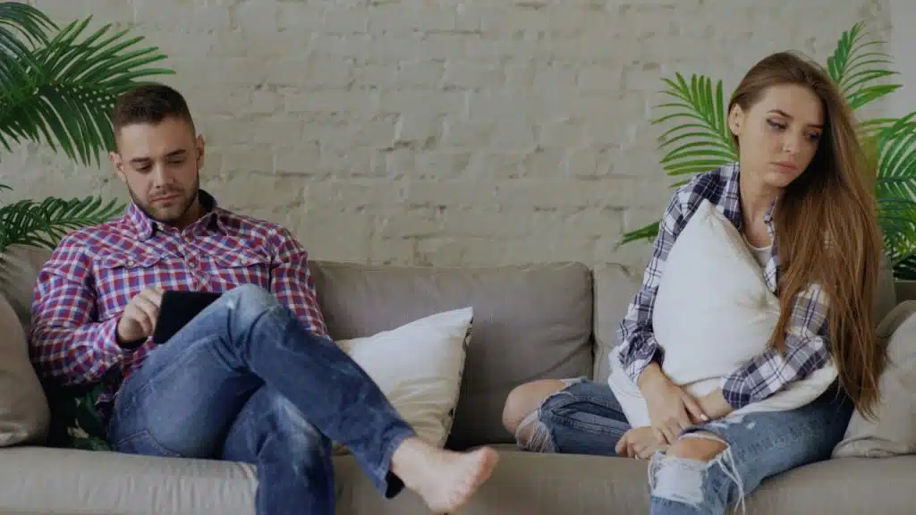 A man using a tablet and a woman holding a pillow sit on a couch.