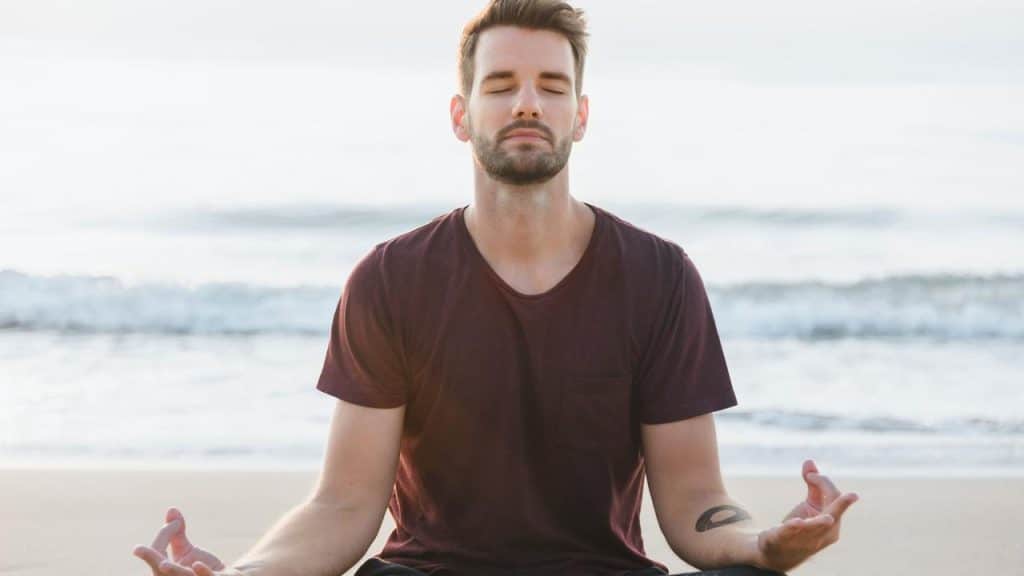 A man meditating on a beach with eyes closed.